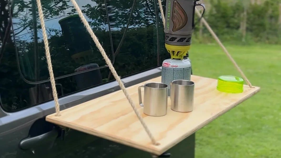 A plywood table hung by rope from the side of a campervan, with a camping stove and two mugs on it