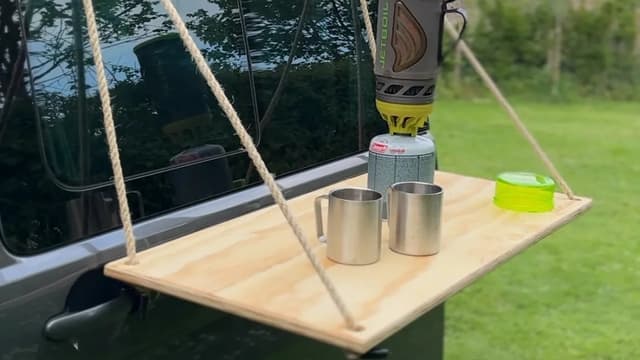 A plywood table hung by rope from the side of a campervan, with a camping stove and two mugs on it
